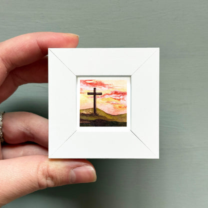 A hand is holding a small white picture frame containing a painting of a cross against a sunset landscape.