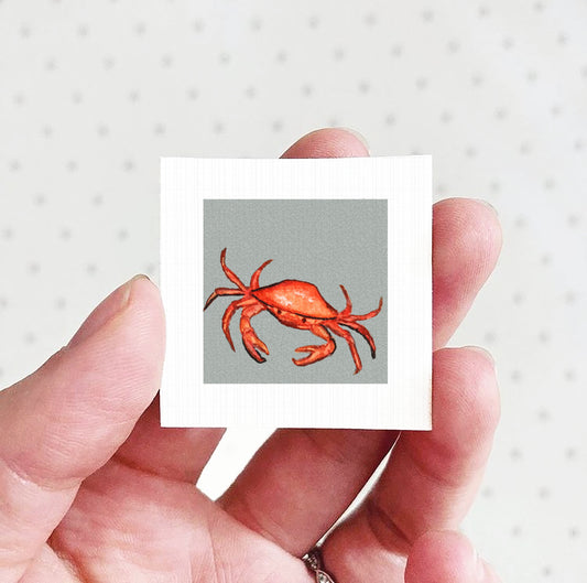 A hand holding a small square picture of a red crab against a gray background.