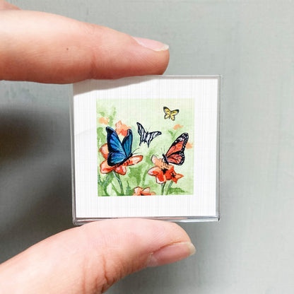 A person's hand is holding a small square picture depicting colorful butterflies on flowers.
