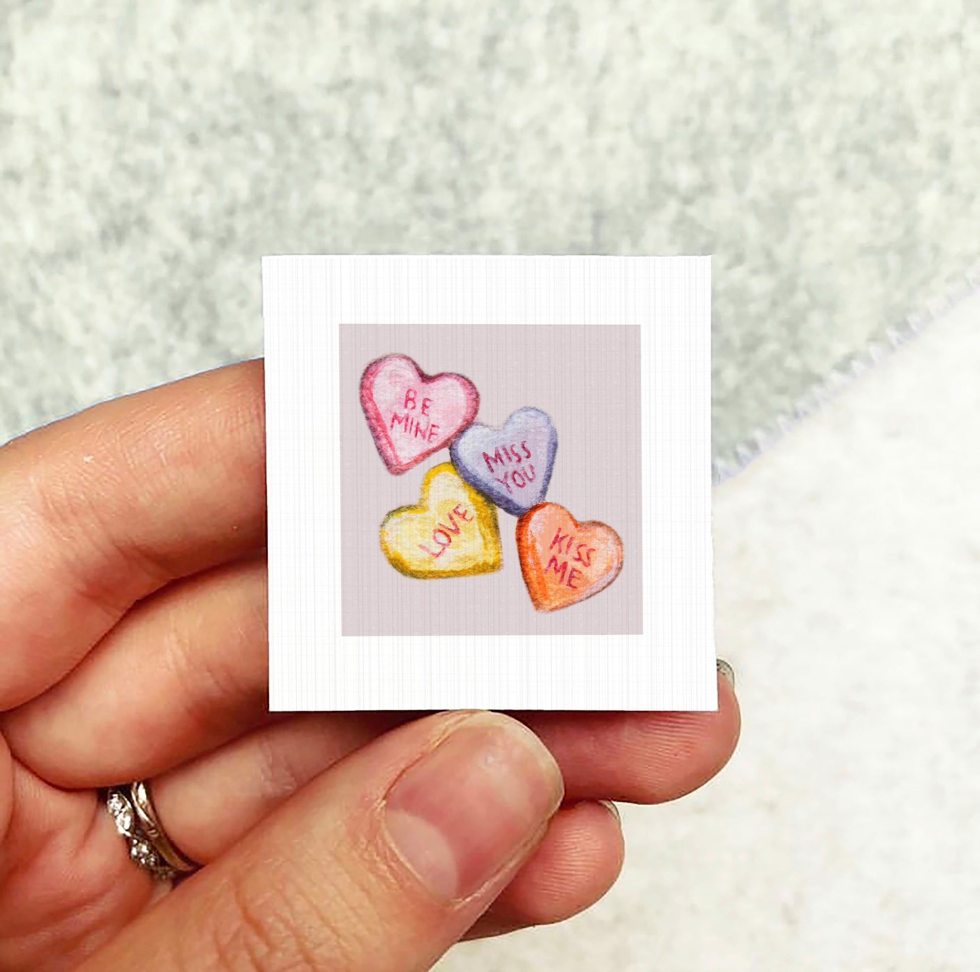 A hand holding a small square card with a heart-shaped candy design on it.