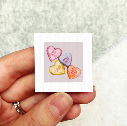 A hand holding a small square card with a heart-shaped candy design on it.