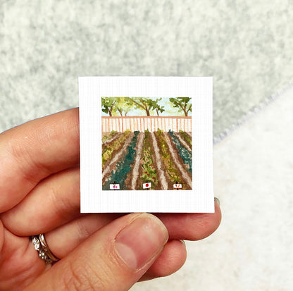 A hand holding a small square artwork depicting a garden scene with rows of plants and a fence.