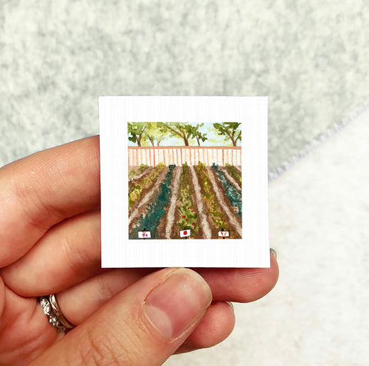 A hand holding a small square artwork depicting a garden scene with rows of plants and a fence.