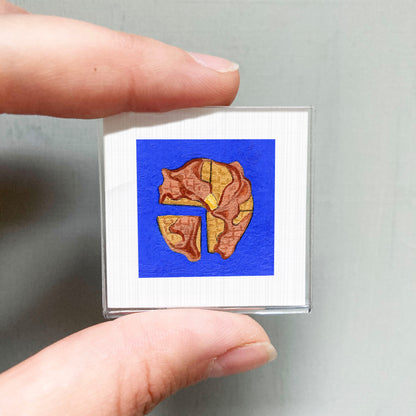 A person's hand is holding a small square with a blue background and a brown and orange design on it.