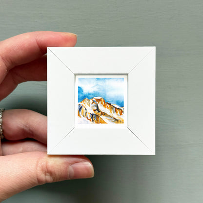 A hand is holding a small white square frame containing a watercolor painting of a mountain landscape.