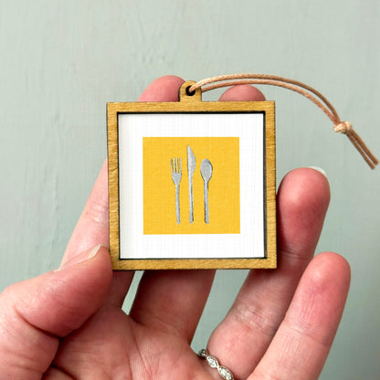 A hand holding a small wooden frame with a picture of a fork, knife, and spoon inside it.