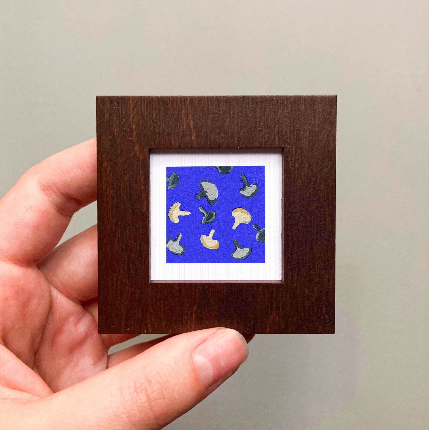 A hand is holding a small square frame containing a vibrant blue background with a pattern of mushrooms.