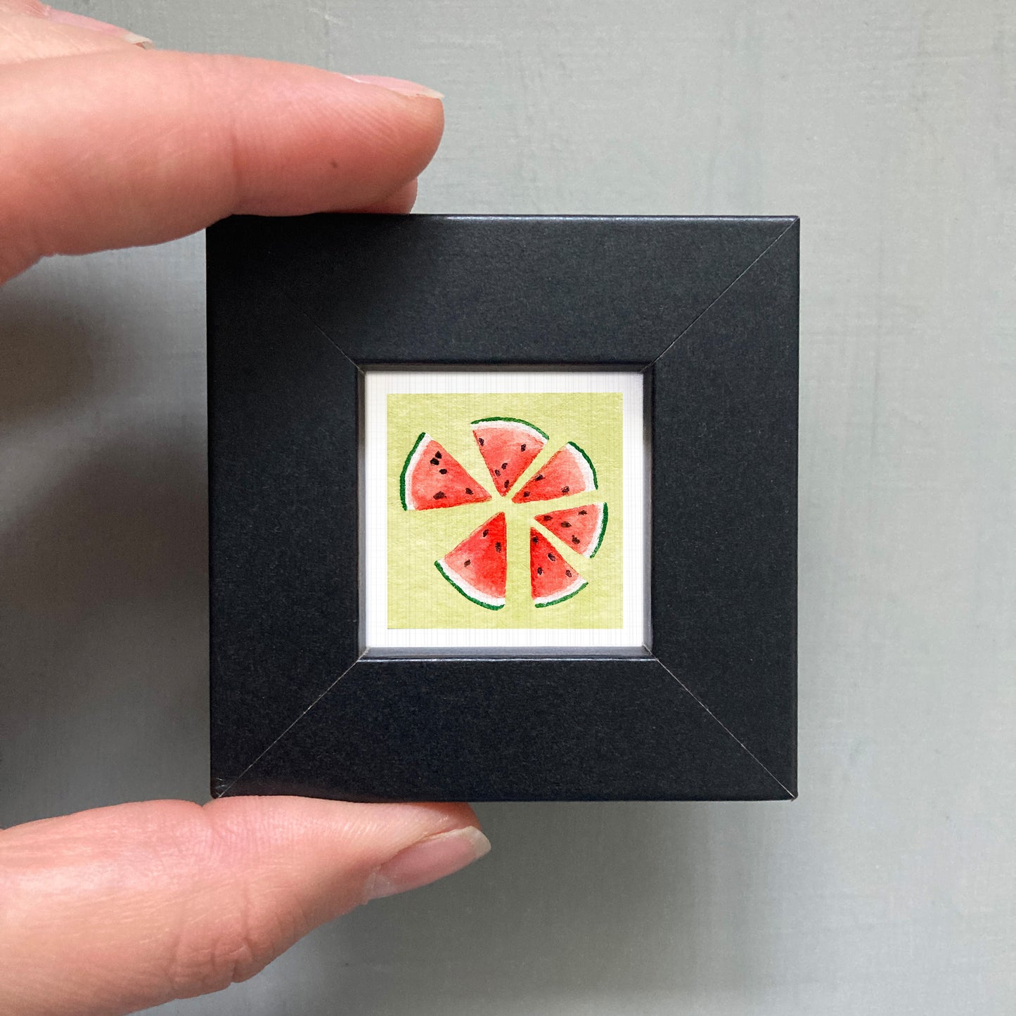 A hand is holding a small square picture frame containing a watermelon slice illustration.