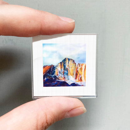 A person's hand is holding a small square picture of a mountain landscape.