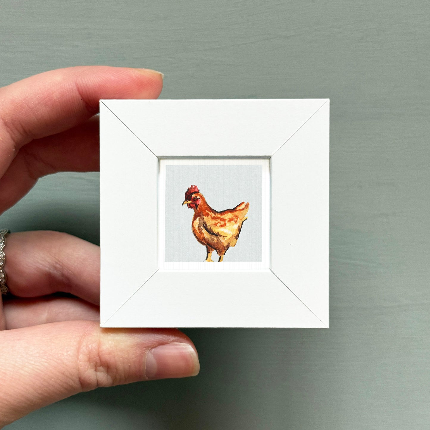 A hand is holding a small white square frame containing a painting of a chicken.