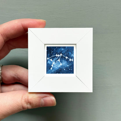 A hand is holding a small white frame containing a framed image of a starry night sky.