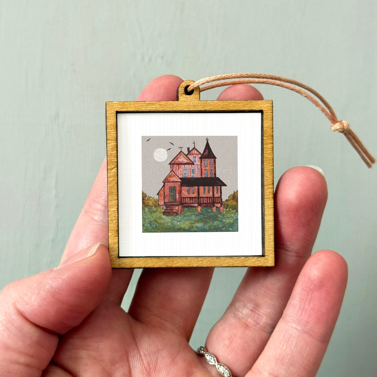 A hand holding a small wooden frame containing a painting of a house with a moon in the background.