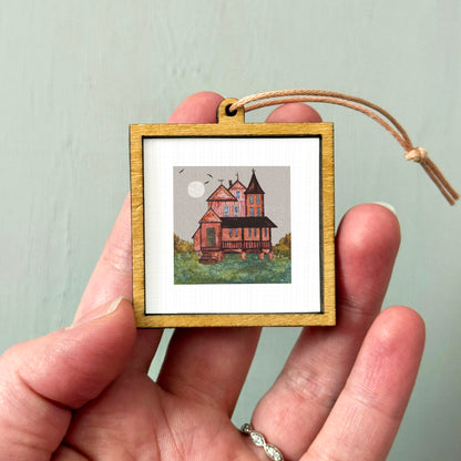 A hand holding a small wooden frame containing a painting of a house with a moon in the background.