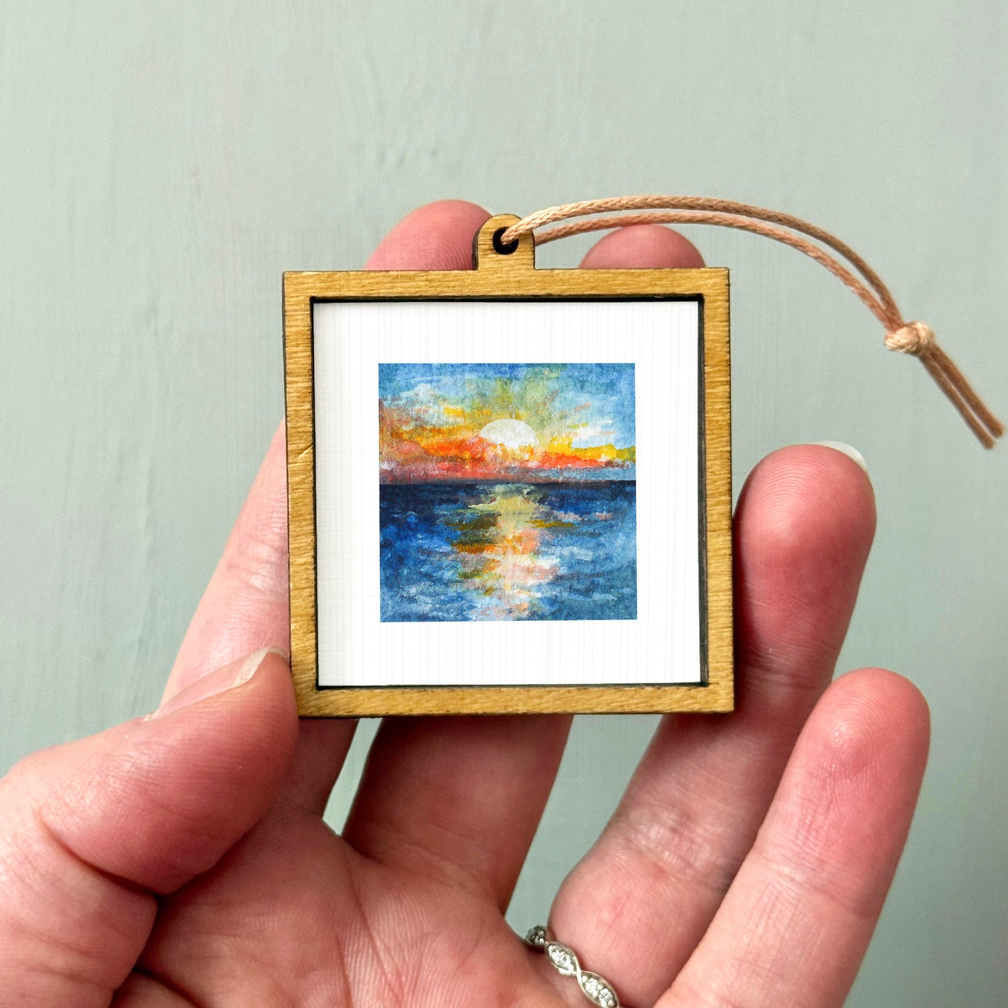 A hand holding a small wooden frame containing a painting of a sunset over a body of water.