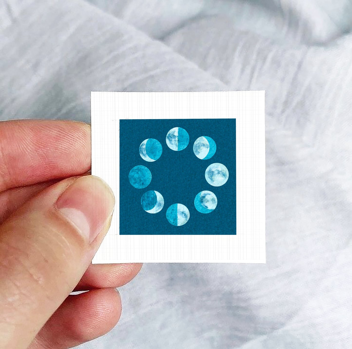 A hand is holding a small square-shaped object with a blue background and a circular arrangement of white and blue circles representing the phases of the moon.