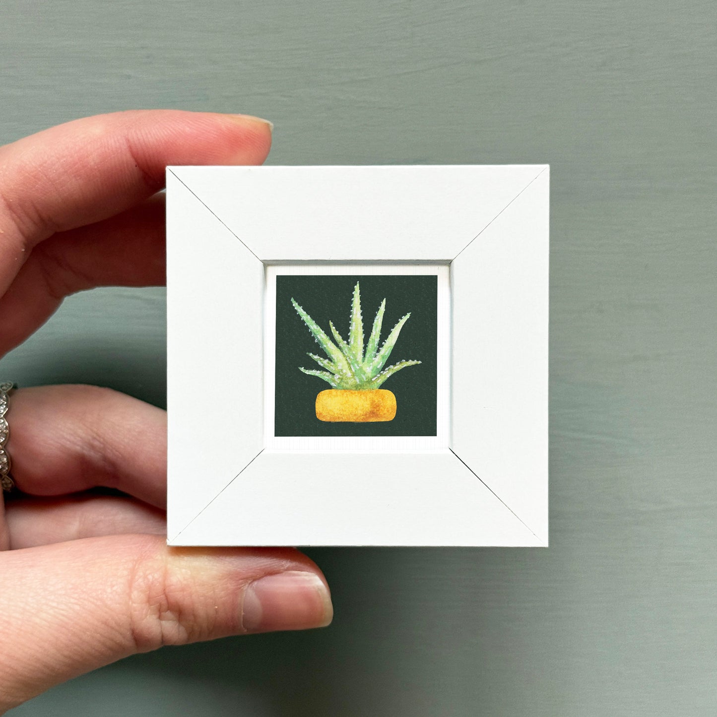 A hand is holding a small white picture frame containing a painting of a cactus plant.
