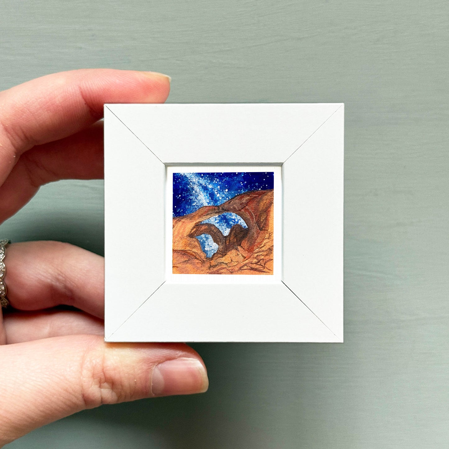 A hand is holding a small white square frame containing a painting of a desert landscape with a starry night sky.