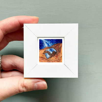 A hand is holding a small white square frame containing a painting of a desert landscape with a starry night sky.