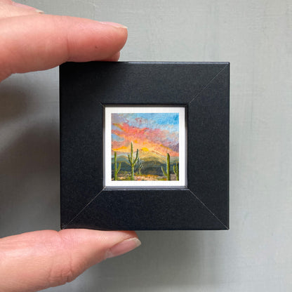 A person's hand holding a small framed painting of a desert landscape with cacti and mountains in the background.