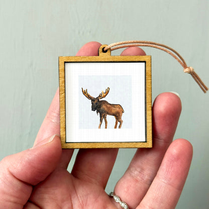 A person's hand holding a small wooden frame containing a picture of a moose.