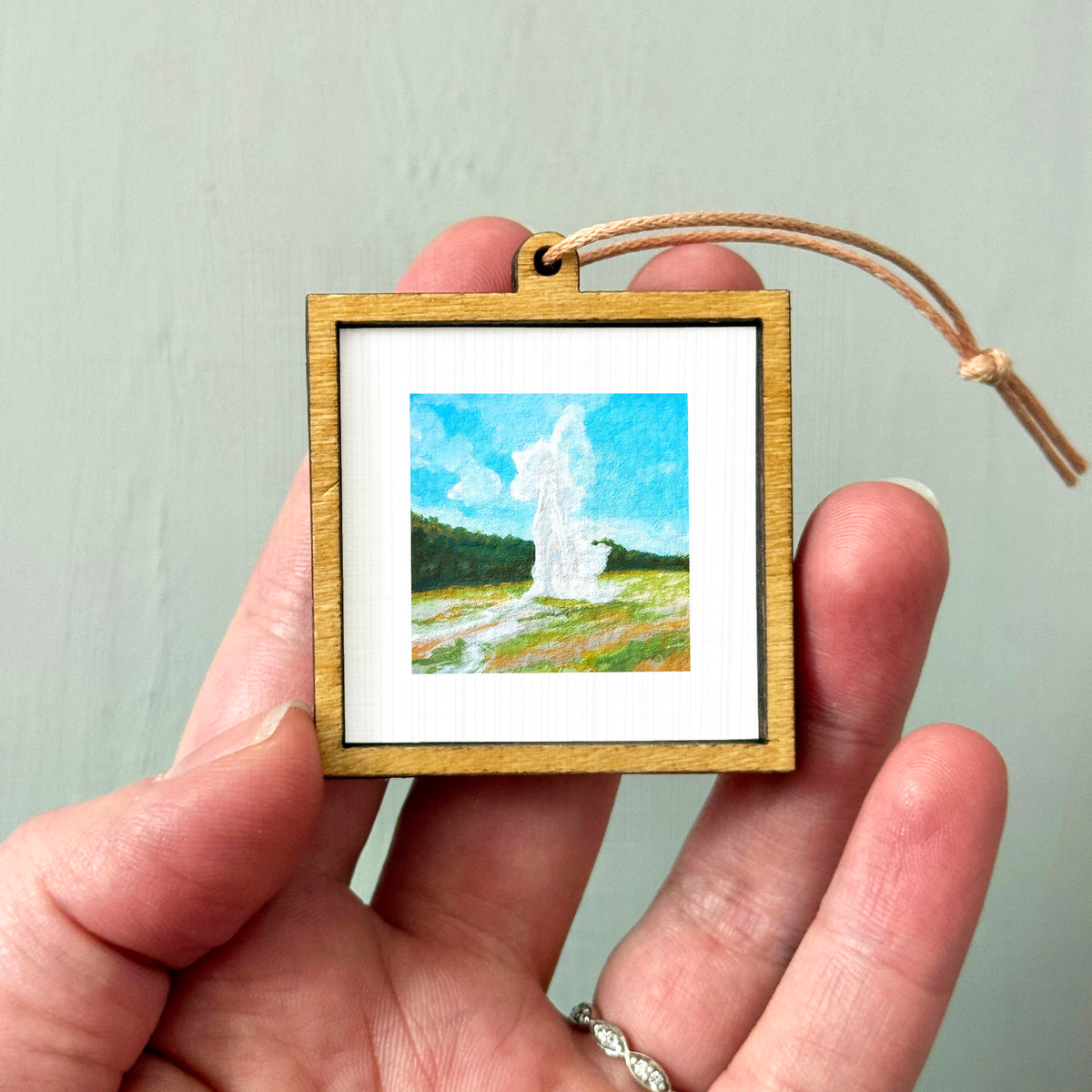 A person's hand holding a small wooden frame containing a painting of a landscape with a tall structure in the background.
