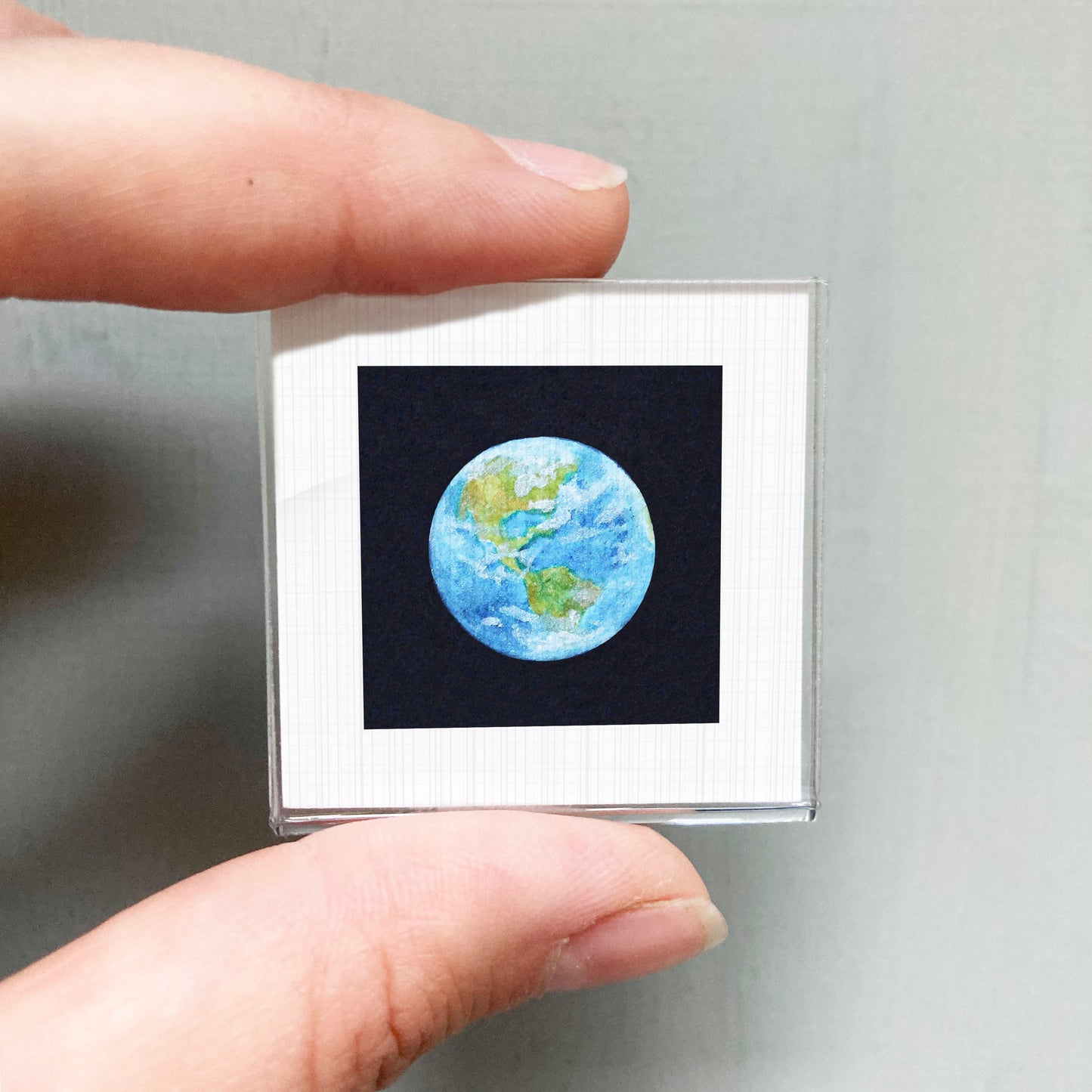 A person's hand holding a small square frame containing a miniature representation of the Earth.