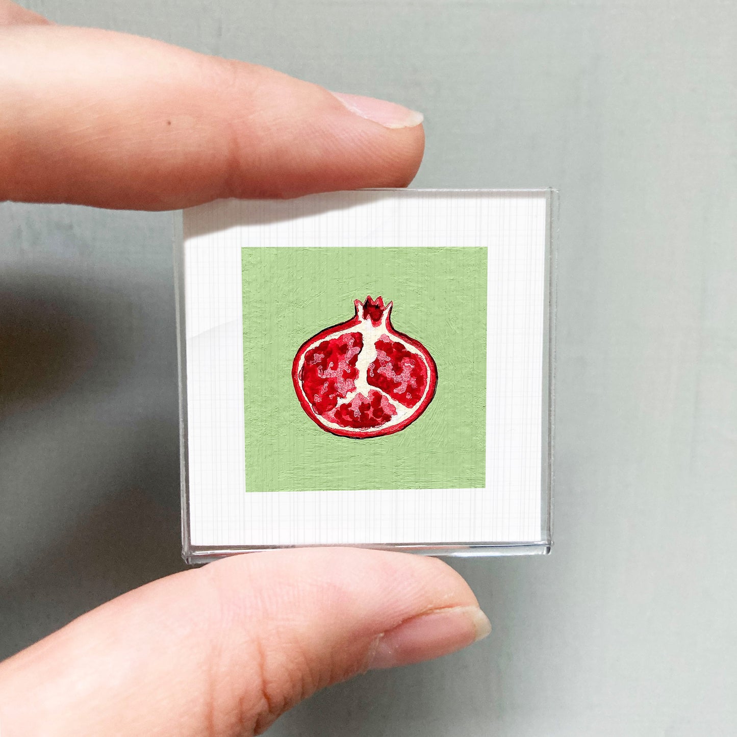 A person's hand holding a small square with a red pomegranate image on a green background.