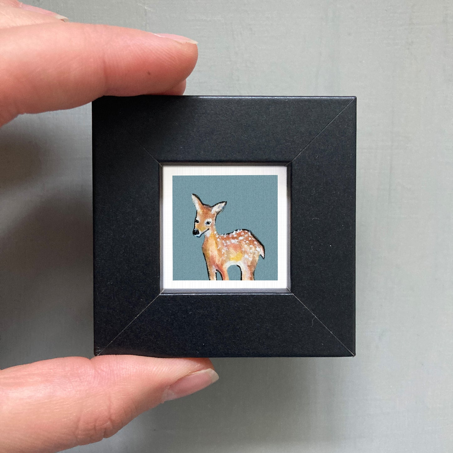 A person's hand holding a small black picture frame containing a painting of a deer.