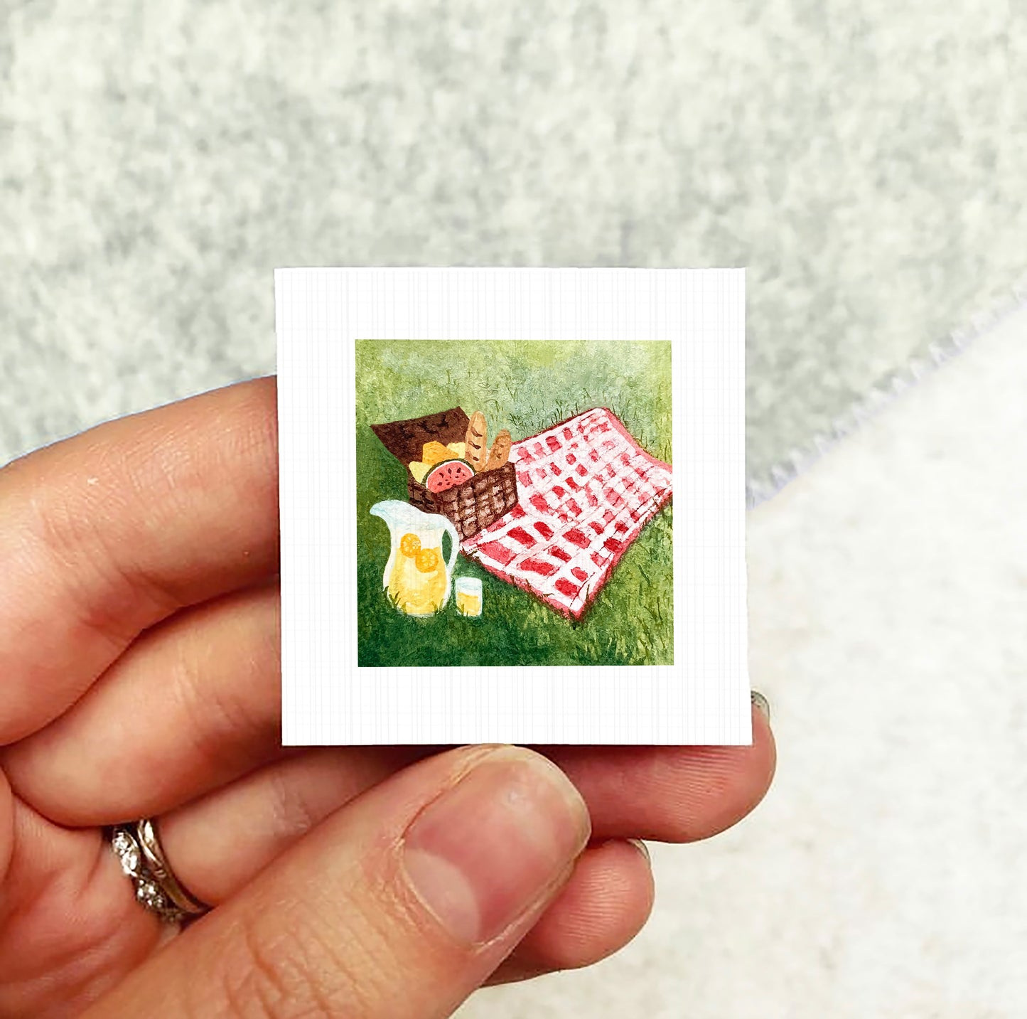 A hand holding a small square picture depicting a picnic scene with a basket, a pitcher, and a red and white checkered blanket on the grass.