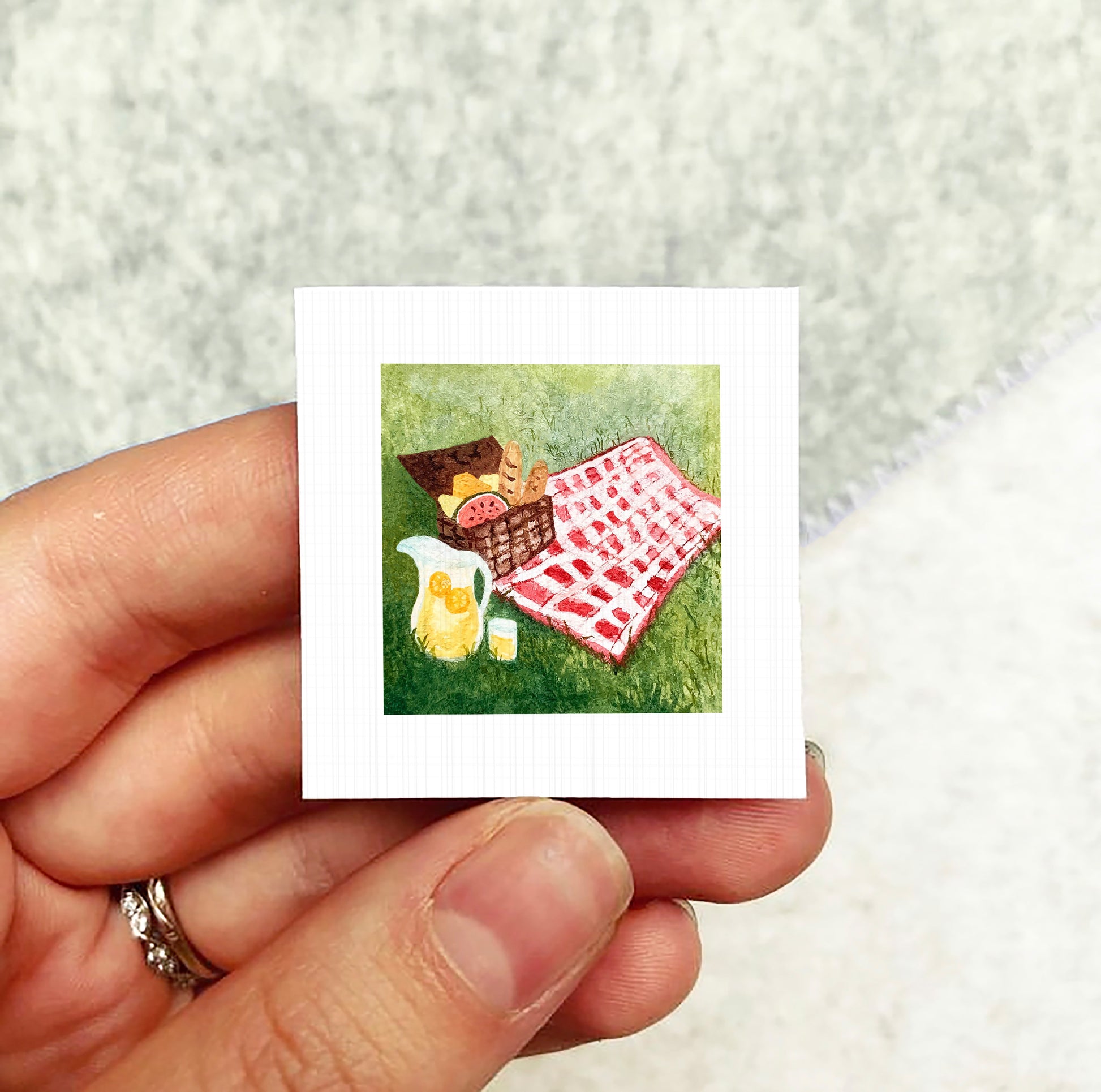 A hand holding a small square picture depicting a picnic scene with a basket, a pitcher, and a red and white checkered blanket on the grass.