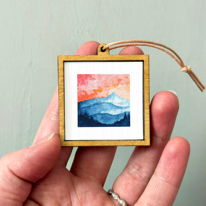 A hand holding a small wooden frame containing a watercolor painting of a mountain landscape.