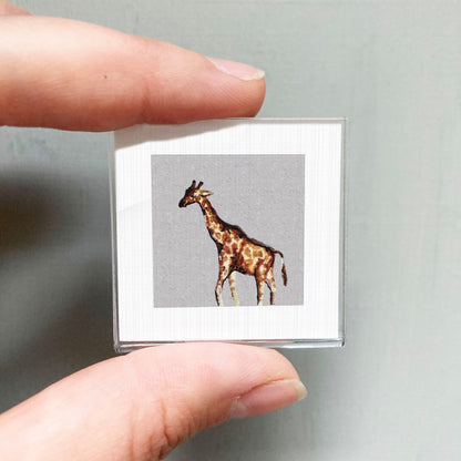 A person's hand is holding a small square picture of a giraffe against a gray background.