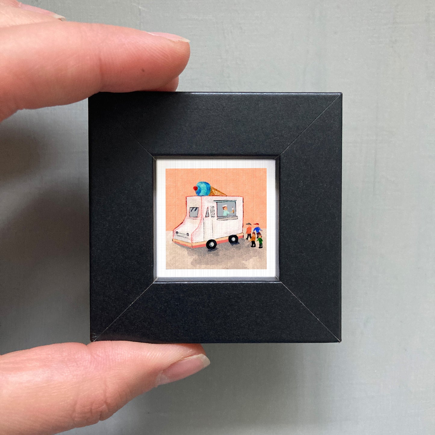 A person's hand is holding a small black frame containing a framed illustration of a white ice cream truck with a blue cone on top.