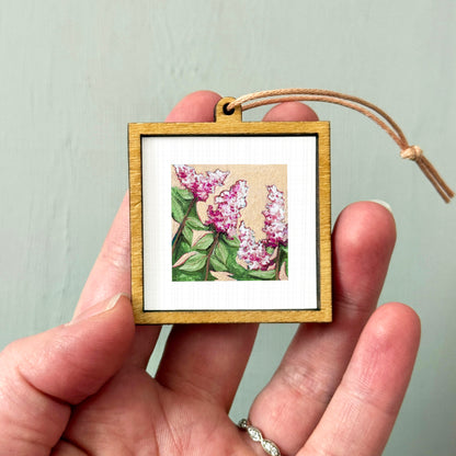 A hand holding a small wooden frame containing a painting of pink flowers.