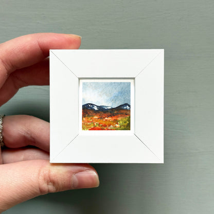 A hand is holding a small white square frame containing a painting of a mountain landscape.
