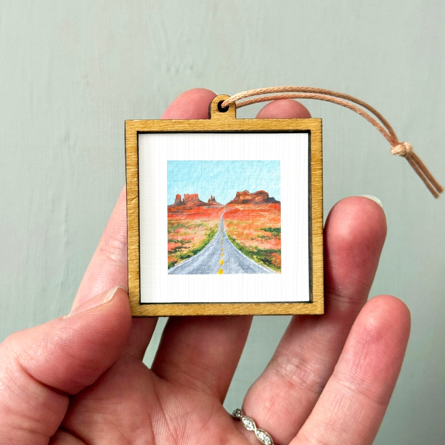 A hand holding a small wooden frame containing a painting of a road leading to a desert landscape.