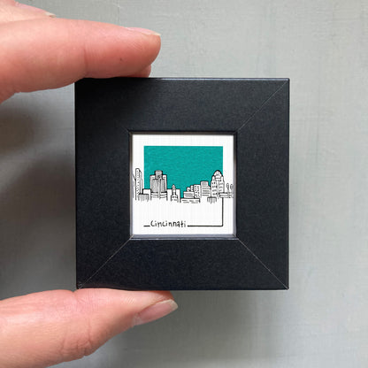 A hand is holding a small black square frame containing a miniature illustration of the Cincinnati skyline.