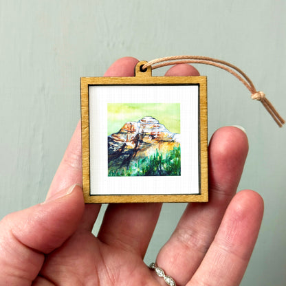 A hand holding a small wooden frame containing a watercolor painting of a mountain landscape.