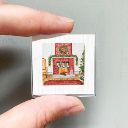 A hand holding a small, square-shaped picture depicting a cozy fireplace scene with a Christmas tree and stockings.