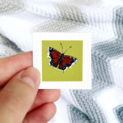 A hand is holding a small square card with a colorful butterfly image on it.