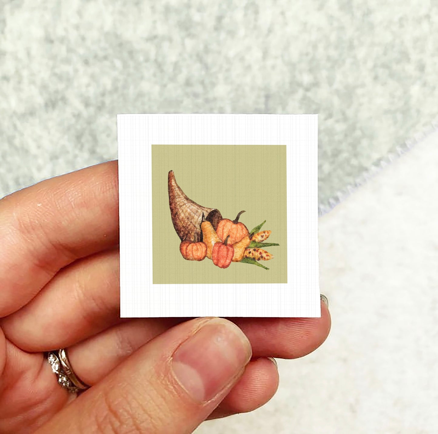 A hand holding a small square card with an illustration of a cornucopia filled with various fall fruits and vegetables.