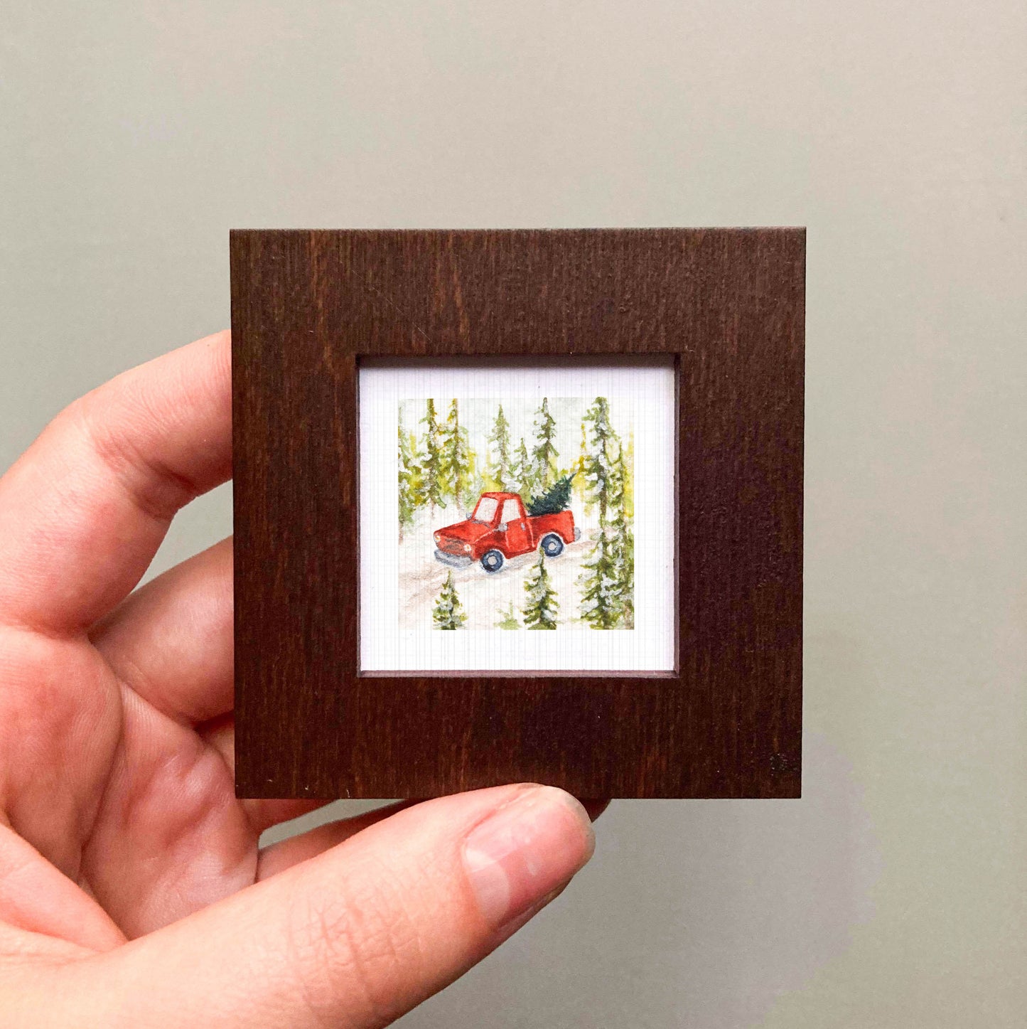 A hand is holding a small, square-shaped picture frame containing a miniature painting of a red truck driving through a snowy forest.