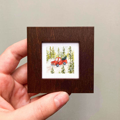 A hand is holding a small, square-shaped picture frame containing a miniature painting of a red truck driving through a snowy forest.