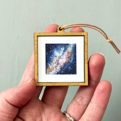 A hand holding a small wooden frame containing a painting of a galaxy.