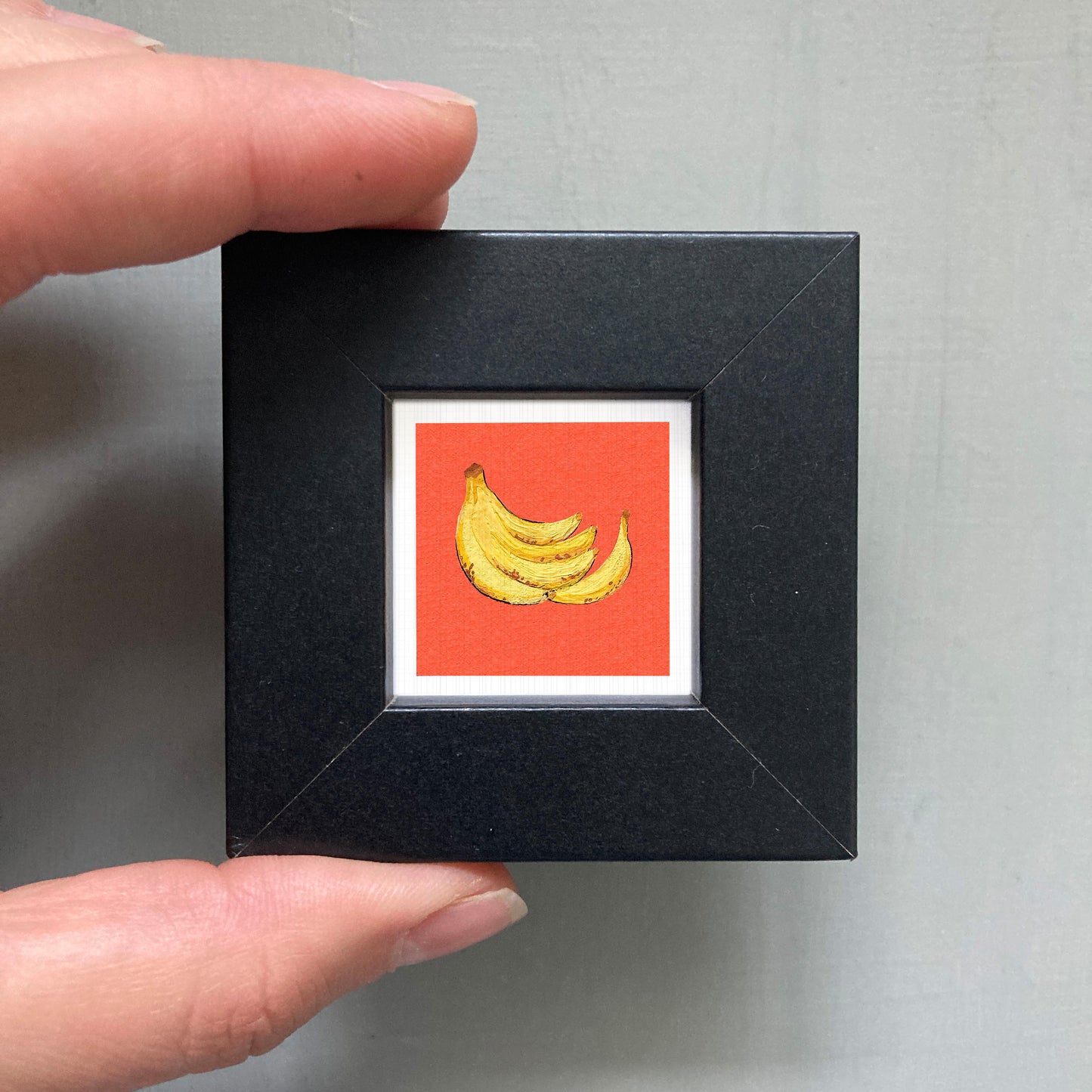 A hand is holding a small black square frame containing a vibrant red square with a yellow banana illustration.