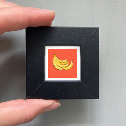 A hand is holding a small black square frame containing a vibrant red square with a yellow banana illustration.