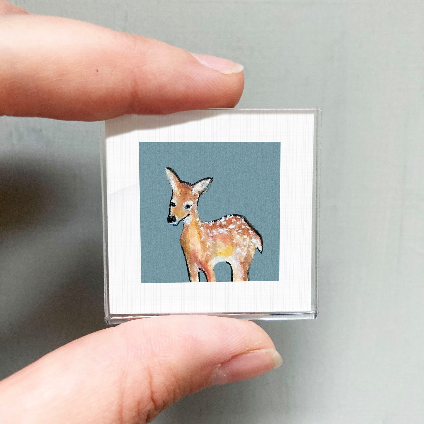 A person's hand is holding a small square picture of a deer against a blue background.