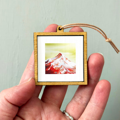 A person's hand holding a small wooden frame containing a framed image of a mountain landscape.