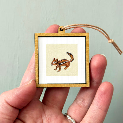 A person's hand holding a small wooden frame containing a picture of a chipmunk.