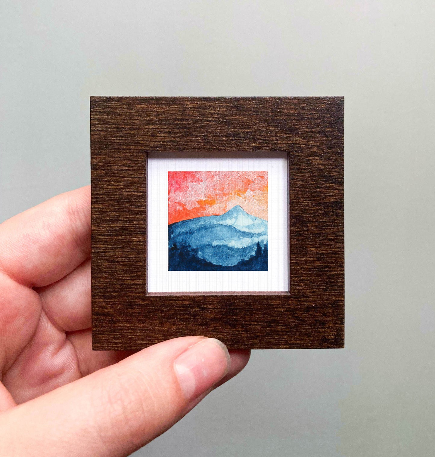 A hand is holding a small, square-shaped wooden frame containing a watercolor painting of a mountain landscape.